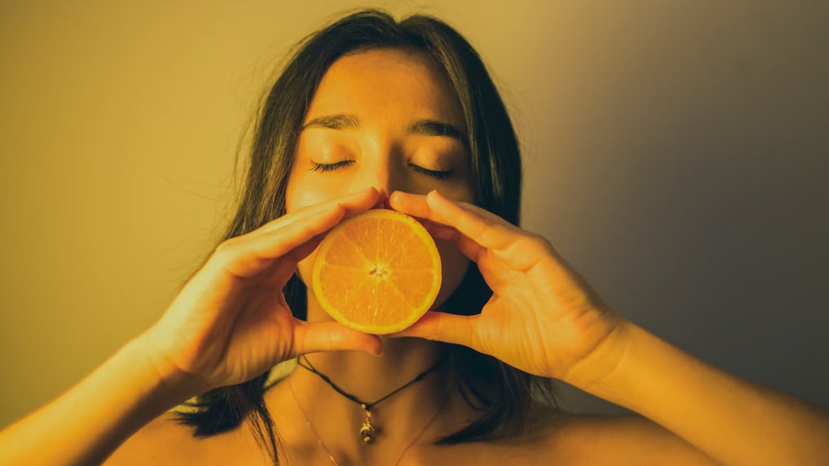 Calm young female with bare shoulders standing with eyes closed against plain wall and covering lips with orange slice